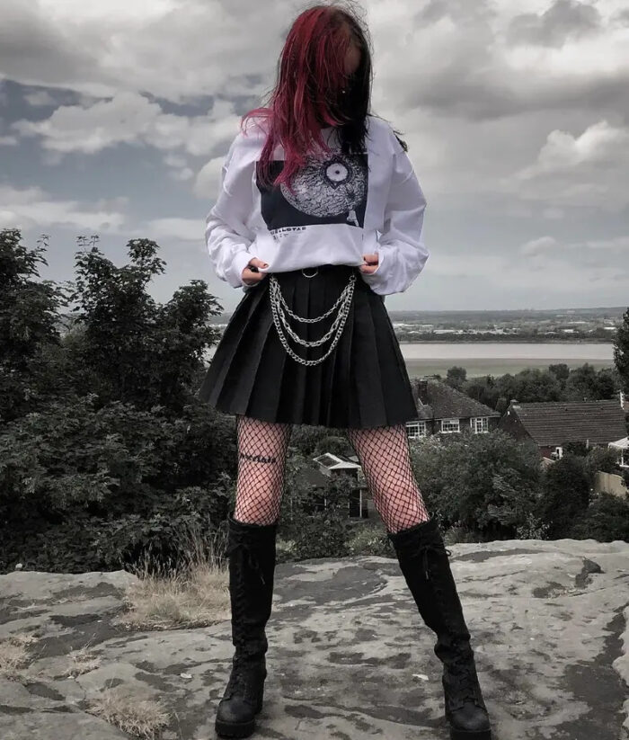 A person with red-tinted hair stands on a rocky surface, wearing a HellStar Discover Sweater, pleated skirt with chain accessories, fishnet stockings, and boots. The background features trees, houses, and a cloudy sky.