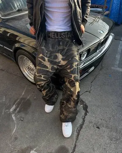 Model wearing Hellstar camo cargo pants with a branded belt standing next to a black car for streetwear fashion.
