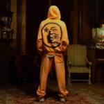 Rear view of a person wearing an orange hoodie with a black flame-haired silhouette graphic and matching sweatpants