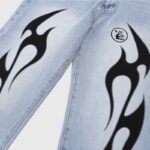 Close-up view of light blue Hellstar denim jeans featuring bold black flame graphics and signature logo detail.