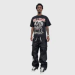 A man standing wearing a black Hellstar Paradise Girls graphic t-shirt and loose black cargo style streetwear pants.