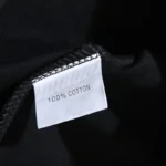 Close-up view of the white fabric tag on a black Hellstar Paradise Girls T-shirt showing 100% cotton material details.