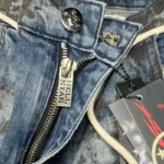 Close-up of the branded silver zipper hardware and metal button on blue vintage washed Hellstar Raw Edge denim jeans.