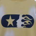 Close-up of the Hellstar Sports yellow sweater featuring a prominent navy blue star and flame pill-shaped graphic logo.