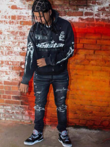 A person wearing Hellstar Studios black racer graphic sweatpants and hoodie against a rustic red brick wall backdrop.
