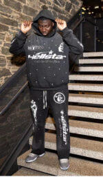 Man wearing Hellstar Studios black Racer Flare sweatpants and matching graphic hoodie standing on stone stairs.