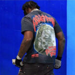 Person seen from the back wearing a black Hellstar Wolf T-shirt featuring a large graphic print against a bright blue wall.