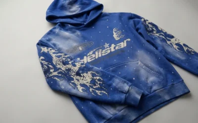 A blue Hellstar hoodie featuring white graphic flame patterns on the sleeves and starry space-inspired print details.